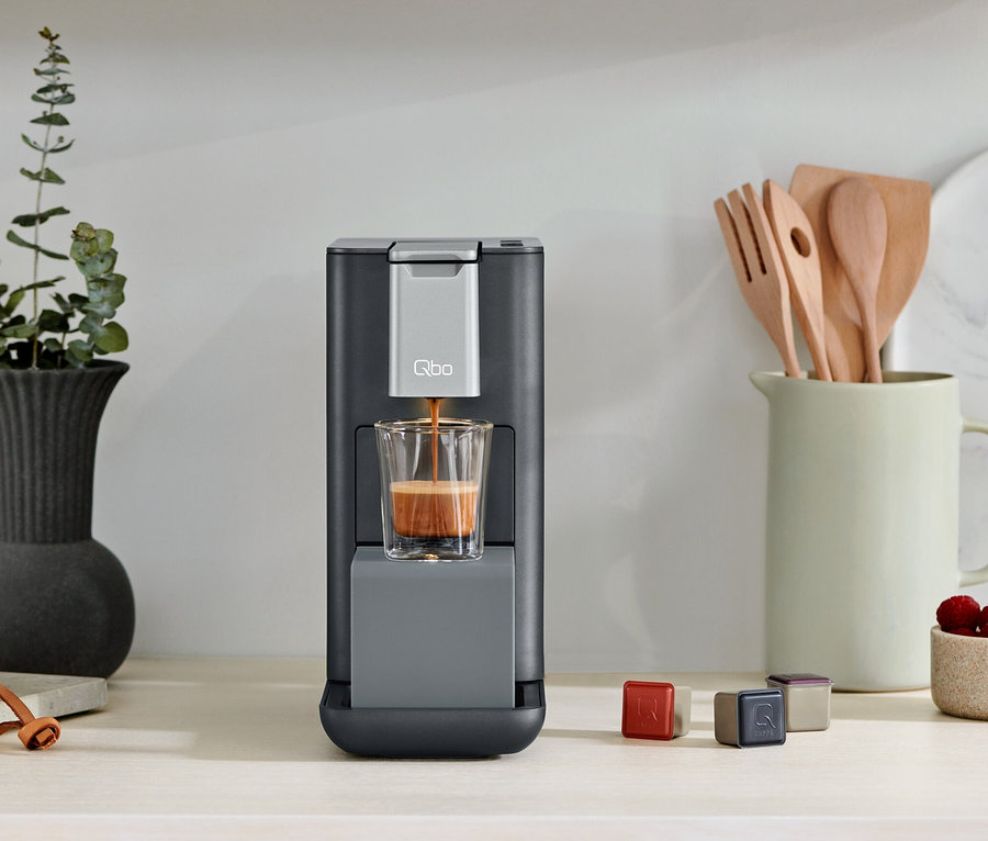 Qbo ESSENTIAL Dark Stone Kaffeemaschine befüllt ein Glas mit Kaffee. Kapseln liegen neben der Kaffeemaschine.