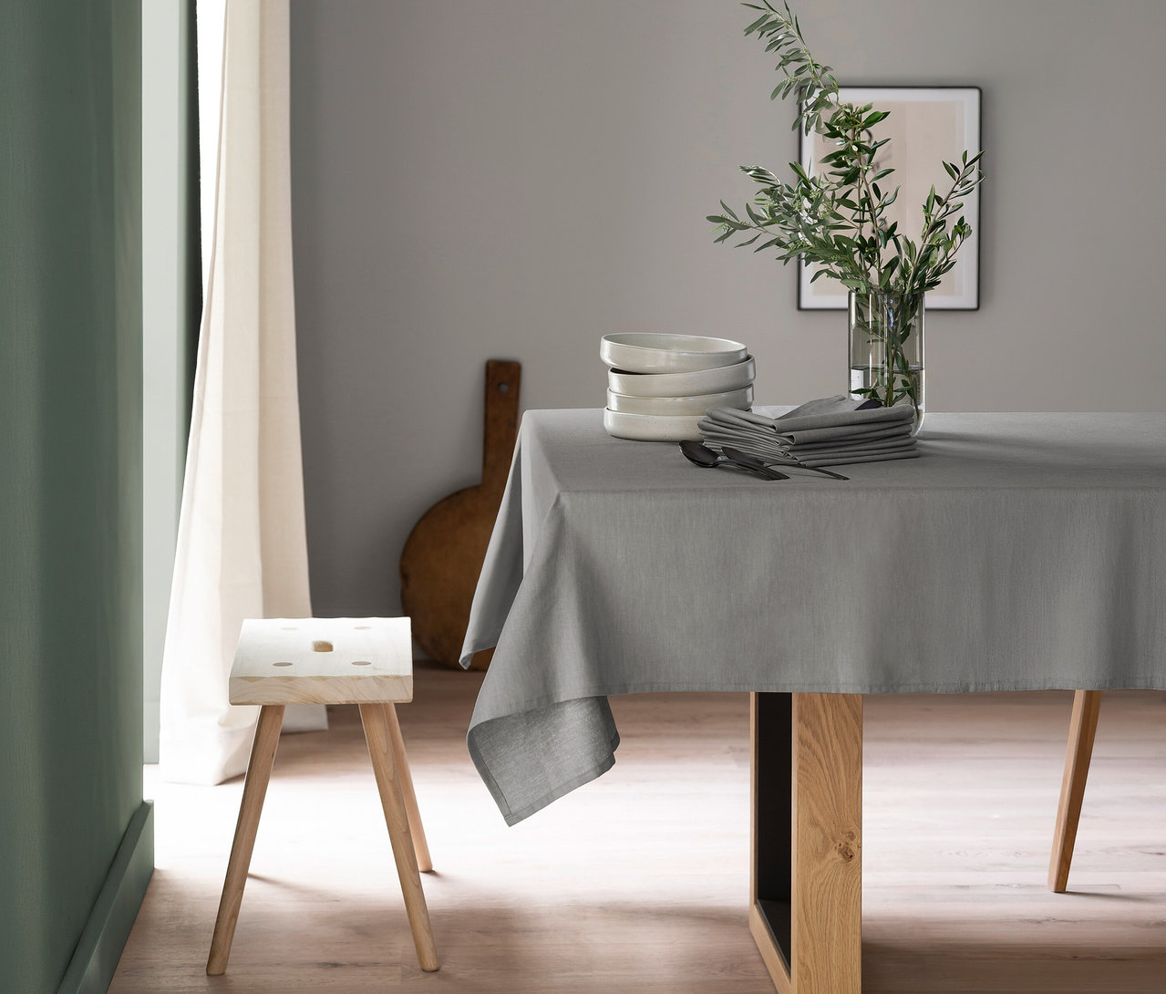Esstisch mit grauer SCHÖNER WOHNEN-Kollektion Tischdecke, darauf weiße Schalen, graue Servietten, Besteck und eine Vase mit grünen Zweigen. Links steht ein Holzschemel.