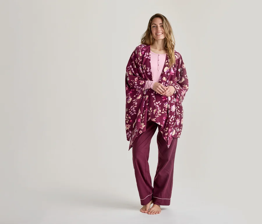 Lächelnde Frau in einem hellrosa Langarmshirt, einer dunkelroten Pyjamahose mit weißer Paspelierung und einem dunkelroten floralen Wickelmantel steht barfuß vor einem weißen Hintergrund.