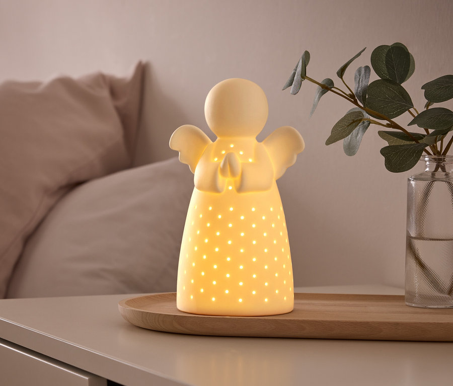 Eine leuchtende, weiße Keramik-Engellampe mit kleinen Perforationen steht auf einem Holztablett auf einem Nachttisch, daneben eine Vase mit einer grünen Pflanze. Im Hintergrund ist ein Bett mit hellrosa Kissen.