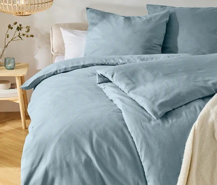 Bett mit hellblauer Bettwäsche und zwei Kissen, einem hellen Kopfteil und einem cremefarbenen Überwurf. Links steht ein Holznachttisch mit Vase und Teelichthalter, darüber hängt eine Holzlampe. Ein runder Teppich liegt auf dem Holzboden.