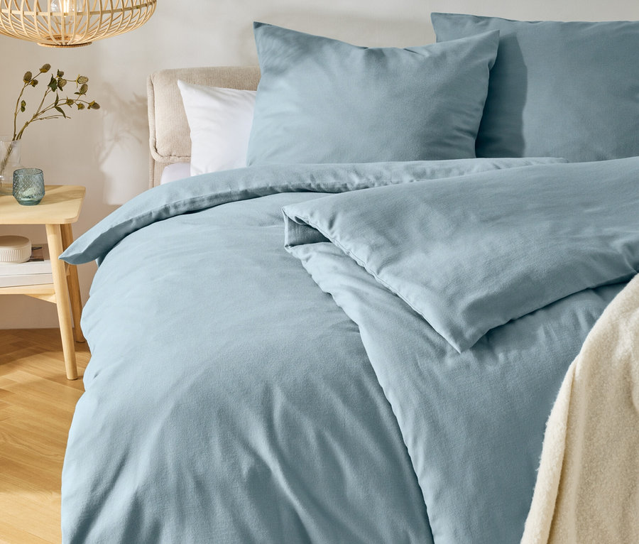 Bett mit hellblauer Bettwäsche und zwei Kissen, einem hellen Kopfteil und einem cremefarbenen Überwurf. Links steht ein Holznachttisch mit Vase und Teelichthalter, darüber hängt eine Holzlampe. Ein runder Teppich liegt auf dem Holzboden.
