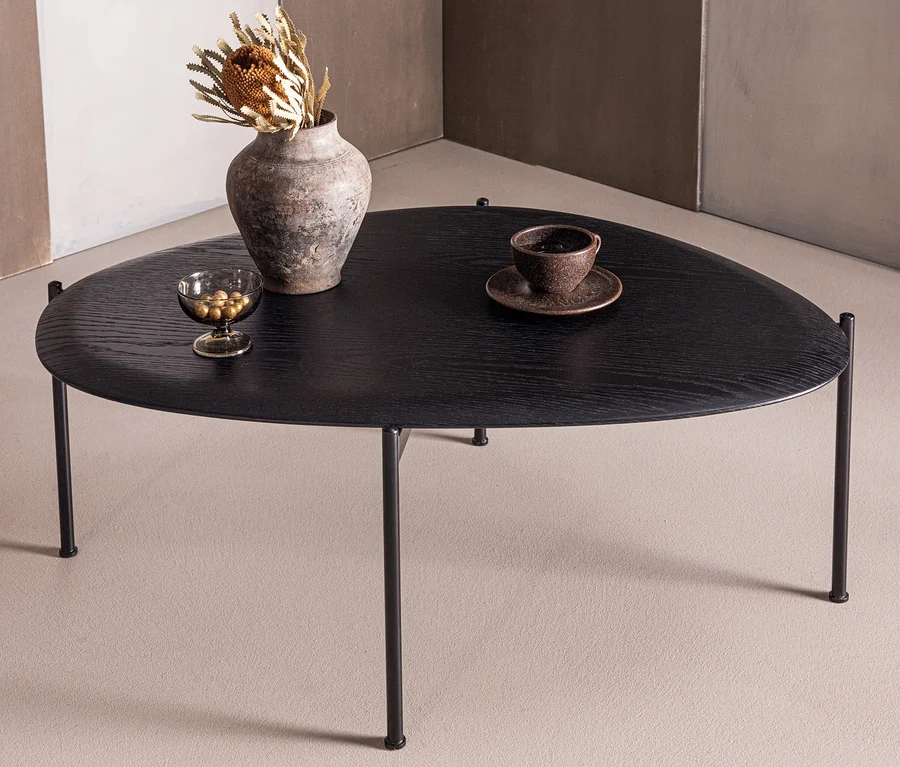 Schwarzer, unregelmäßig geformter Couchtisch mit Metallbeinen. Darauf eine Vase mit Trockenblumen, eine Schale mit goldenen Kugeln und eine Tasse mit Untertasse.