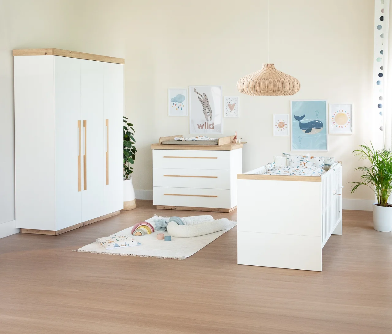 Ein helles Babyzimmer mit einem weißen Roba 2-teiliges Kinderzimmer-Set »Lumo«, bestehend aus einem Kleiderschrank und einer Wickelkommode. Daneben steht ein passendes Gitterbett, ein Teppich mit Spielzeug und Wandbilder mit Tiermotiven.