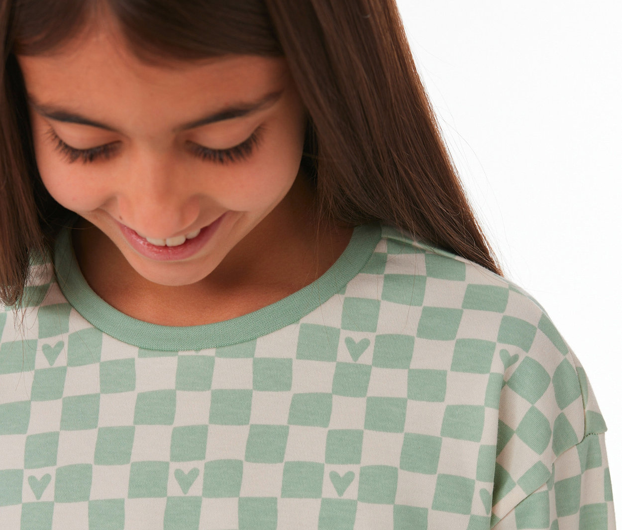 Lächelndes Mädchen blickt nach unten, trägt ein SCHIESSER Kinder-Interlock-Pyjama-Oberteil mit hellgrünem Karomuster und Herzchen.