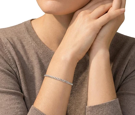 Nahaufnahme einer Person in braunem Strickpullover, die ein Amor 925 Silber Panzer-Armband am linken Handgelenk trägt. Die Hände sind gefaltet.