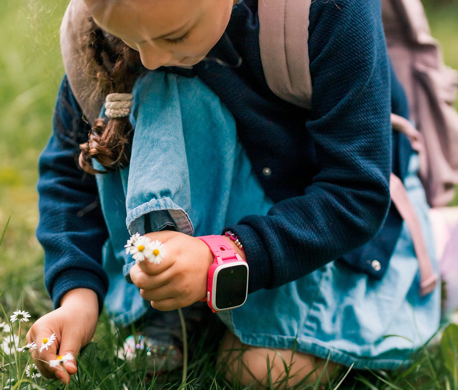 Ein Kind kniet im Gras, pflückt Gänseblümchen und trägt eine rosa Xplora XGO3 Kids Watch.