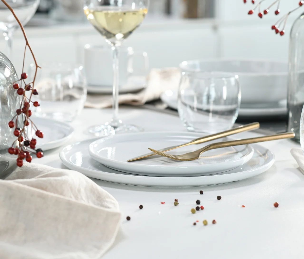 Nahaufnahme eines gedeckten Tisches mit zwei weissen Tellern des creaTable Service »Session White«, goldenem Besteck, einem Weinglas und einer Serviette. Rote Beeren und Pfefferkörner liegen verstreut.
