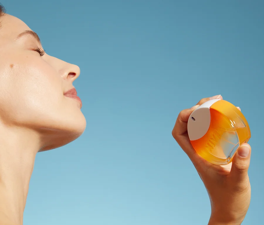 Frau mit geschlossenen Augen und zurückgelegtem Kopf, die eine orange-weiße NIVEA Sprühflasche vor blauem Himmel hält.