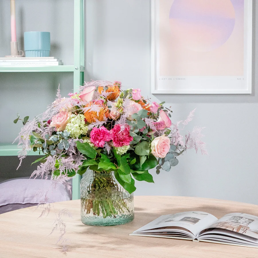 Strauß aus Rosen, Nelken und Proteen in einer durchsichtigen Vase auf einem Tisch neben einem offenen Buch.