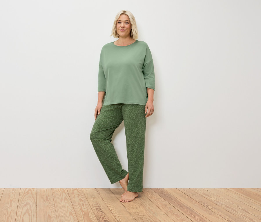 Blonde Frau im grünen Pyjama steht auf Holzboden vor einer weißen Wand.