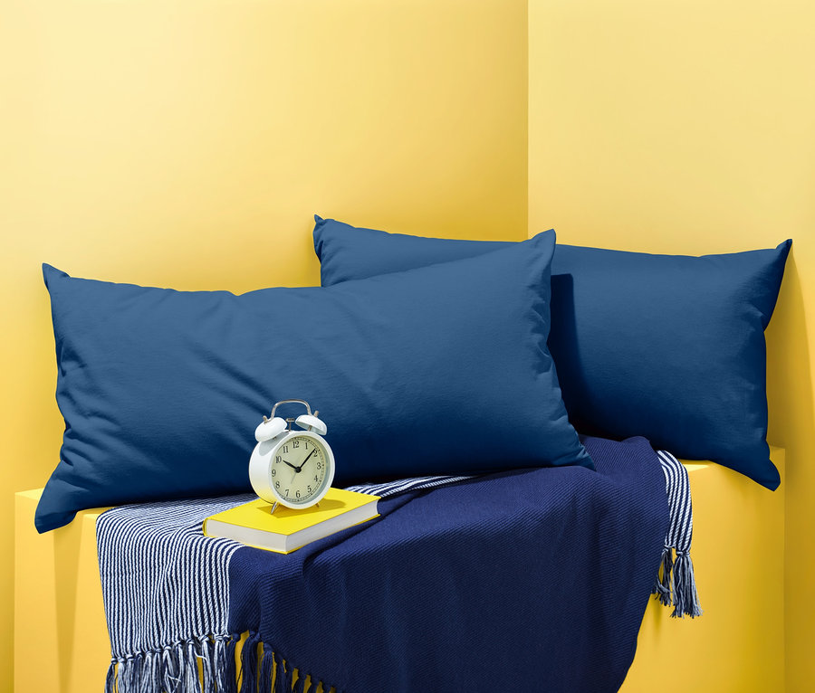 Zwei blaue Jersey-Kopfkissenbezüge liegen auf einer blauen Decke mit einem Buch und einem Wecker.