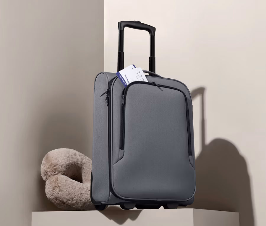 Ein grauer Kabinentrolley steht neben einem beige farbenen Reisekissen.