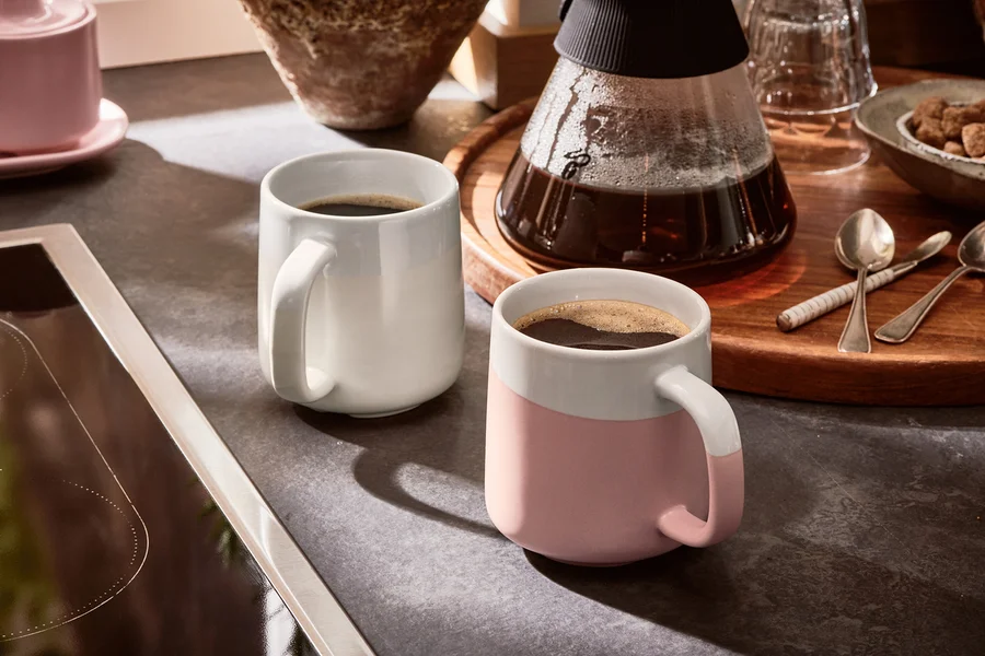 Zwei Kaffeetassen stehen neben einer Kaffeemaschine auf einem Tisch.