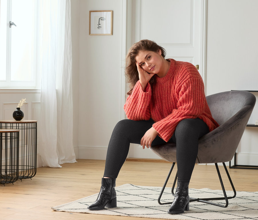 Frau sitzt im Sessel und trägt einen grob gestrickten Pullover, gefütterte Leggings im Jeans-Look und schwarze Leder-Stiefeletten.