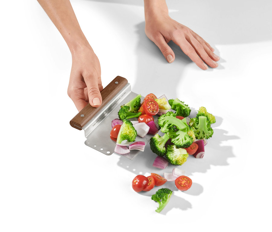 Zwei Hände halten einen 2-in-1-Teigschneider mit geschnittenem Brokkoli, Tomaten und Zwiebeln auf einem weißen Hintergrund.