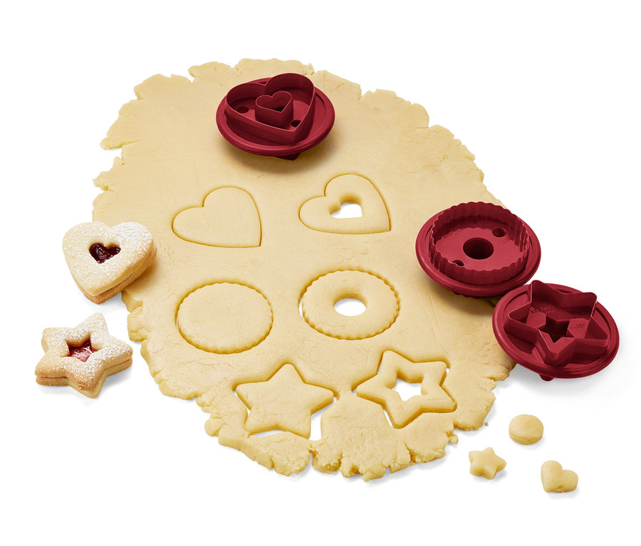 Auf einem Teigroller liegen 3 Linzer Plätzchenausstecher und ausgestochene Teigstücke.