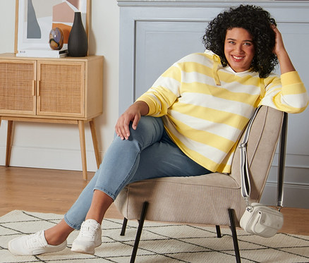 Eine Frau sitzt in einem Sessel und trägt ein gelb-cremefarbenes, gestreiftes Kapuzen-Sweatshirt, Treggings, Veloursleder-Sneaker und eine Umhängetasche.
