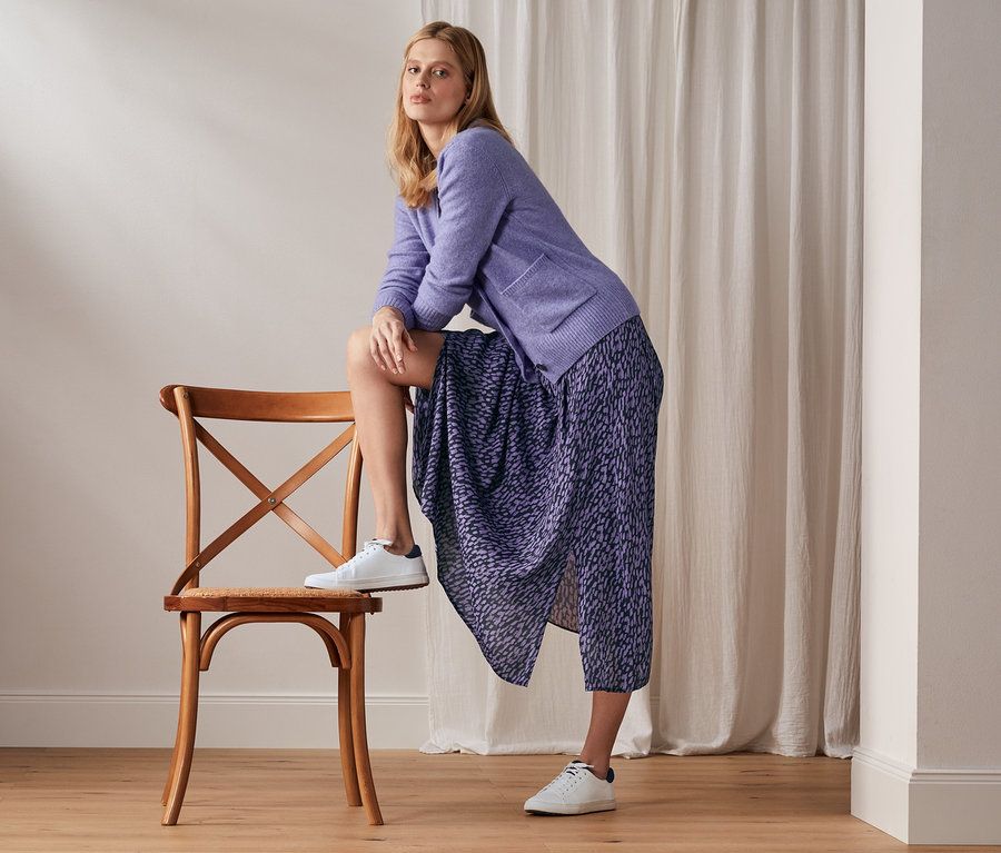 Frau posiert mit einem Bein auf einem Stuhl, trägt einen Rock mit Print, eine lila Strickjacke und weiße Turnschuhe.