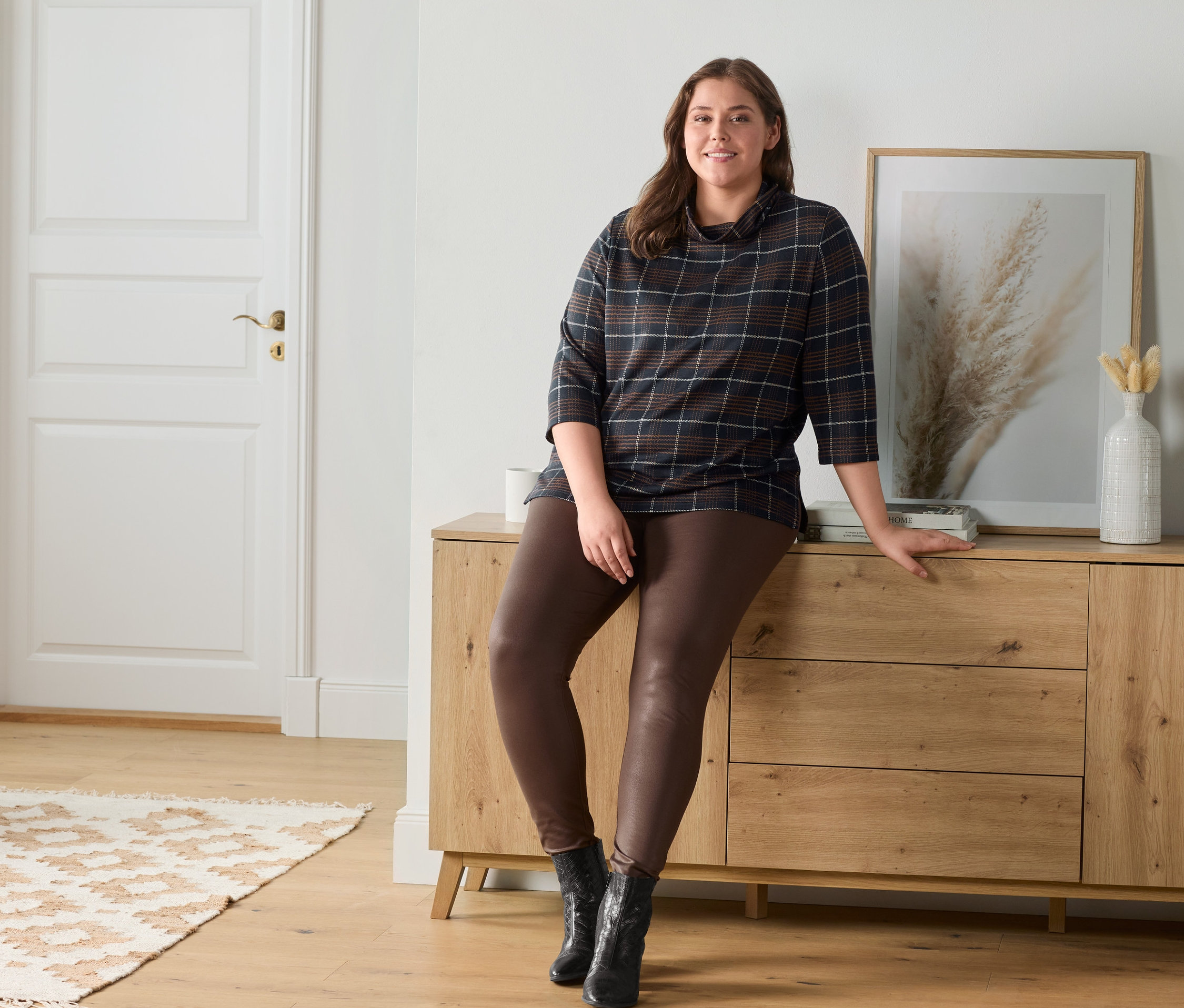 Eine Frau sitzt auf einer Holzkommode und trägt beschichtete Leggings und ein kariertes Sweatshirt.