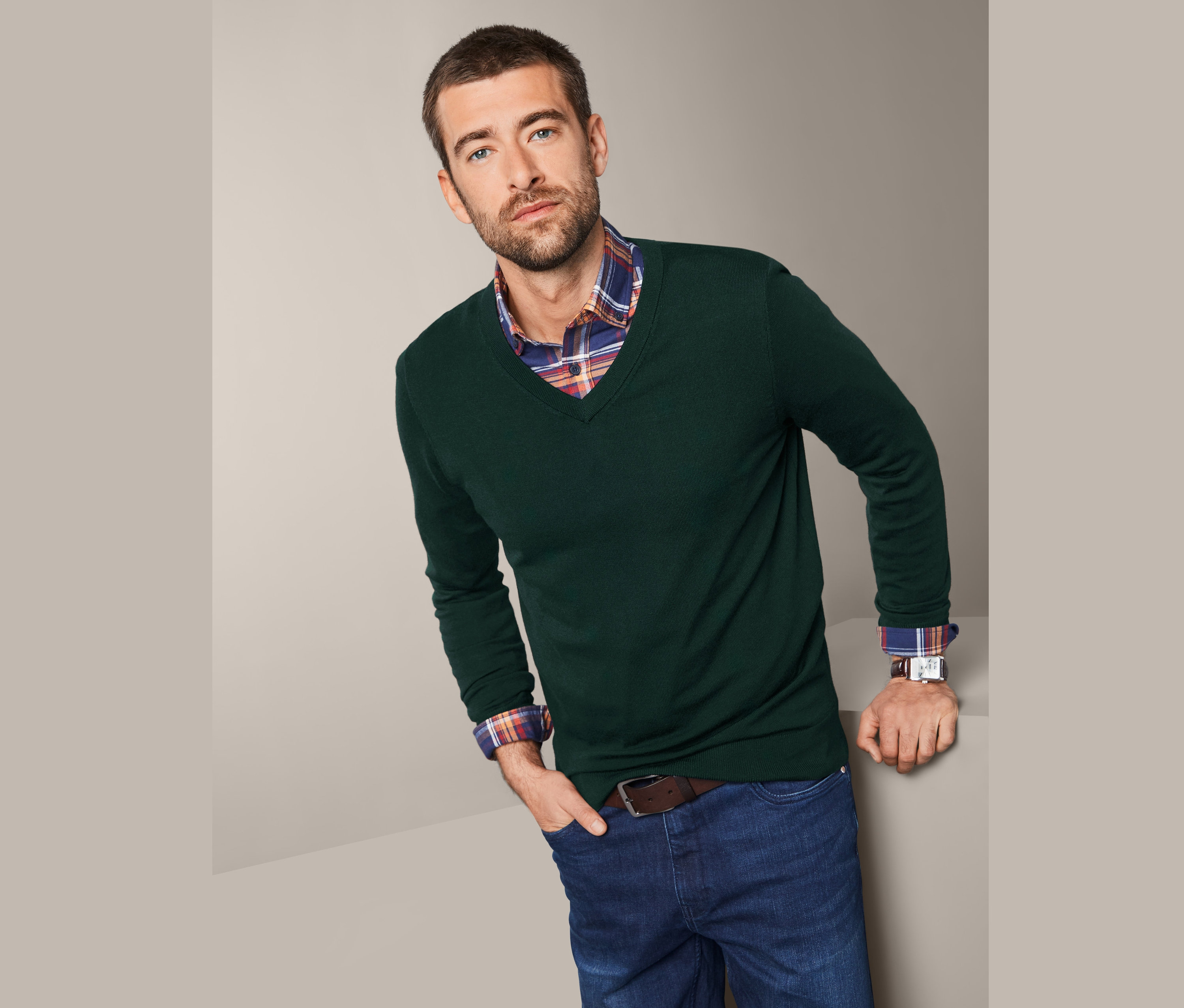 Mann, der sich an eine Wand lehnt, trägt einen grünen Merino-Pullover und Jeans.