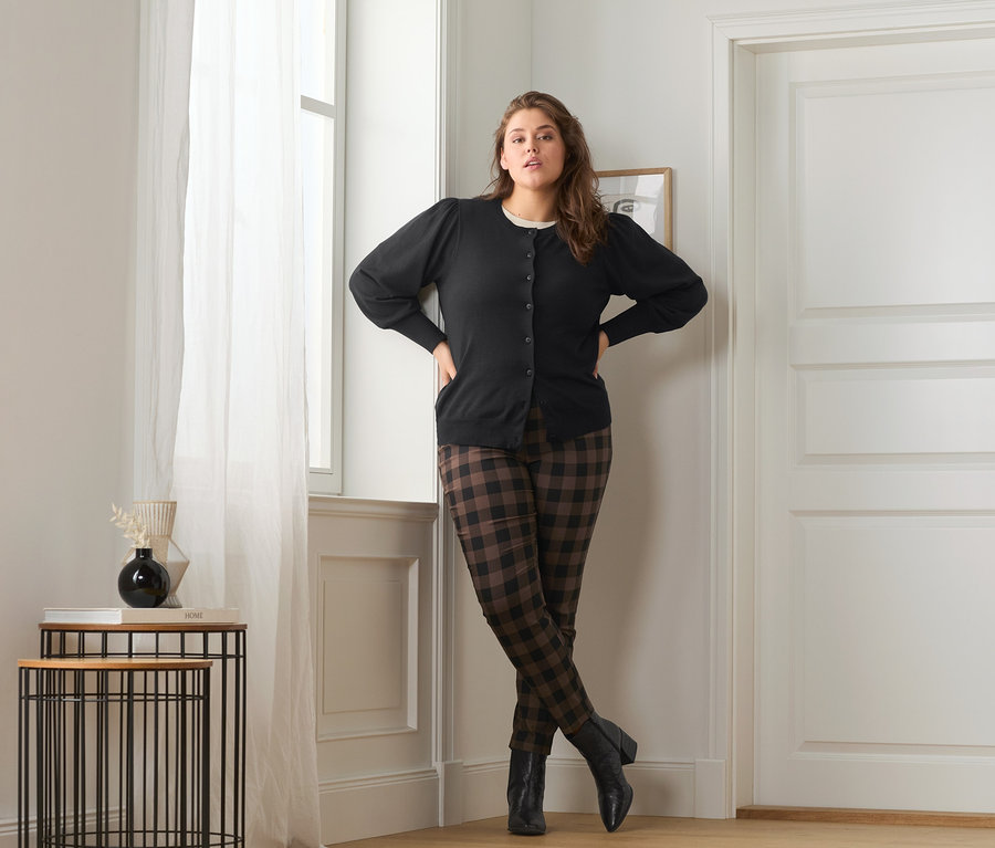 Frau posiert in Feinstrickjacke mit Puffärmeln, karierter Stretchhose und Leder-Stiefeletten.