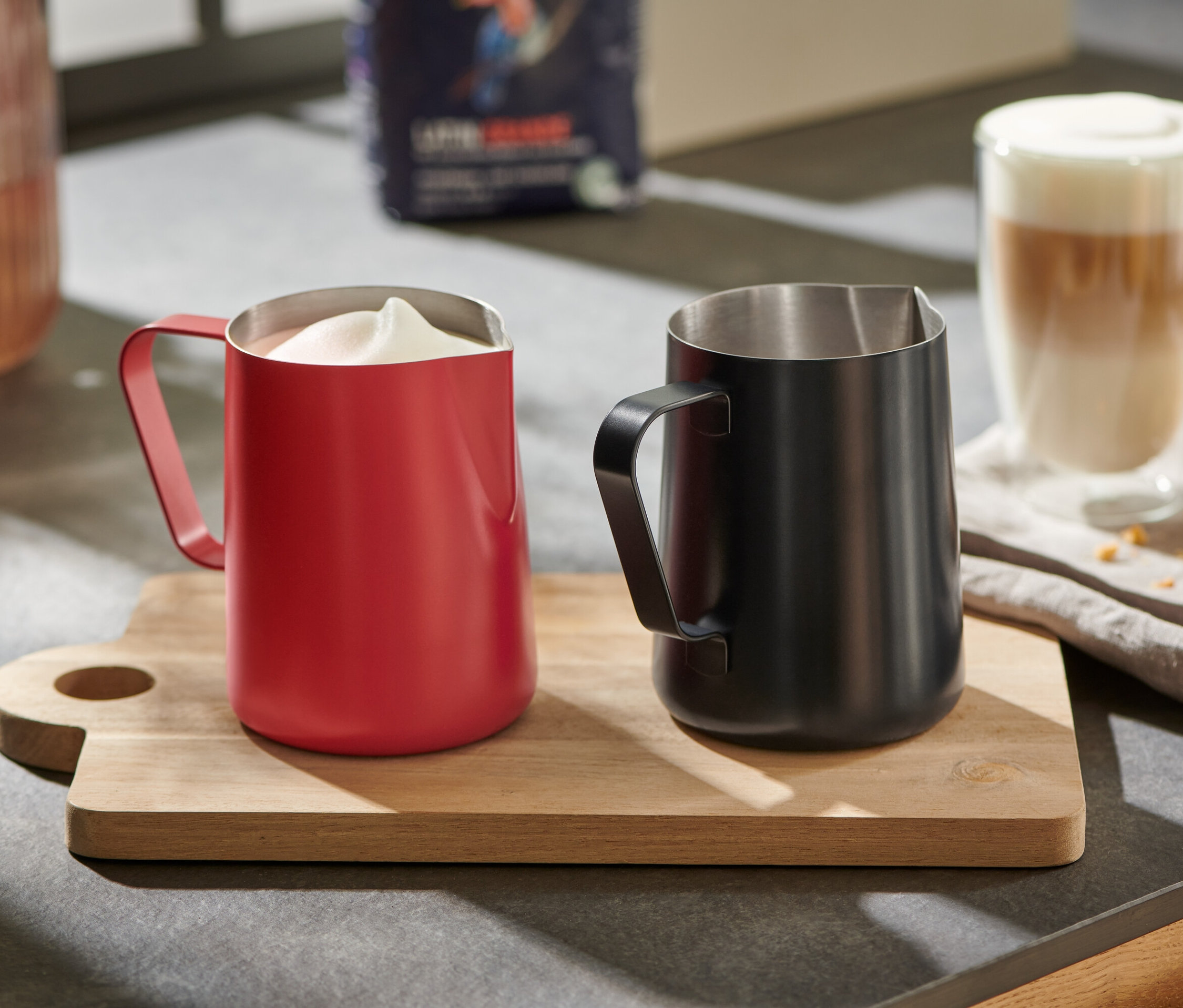 Ein rotes Milchkännchen mit aufgeschäumter Milch und ein schwarzes Milchkännchen auf einem Holzbrett.