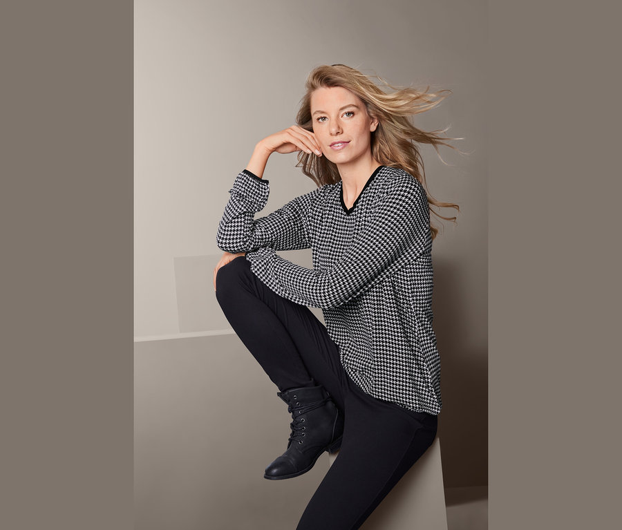 Blonde Frau sitzt und trägt ein bedrucktes Blusenshirt.