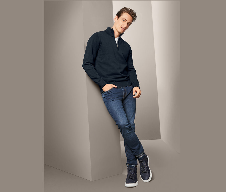 Mann lehnt an Wand, trägt dunkelblauen Grobstrick-Troyer, Jeans und Turnschuhe.