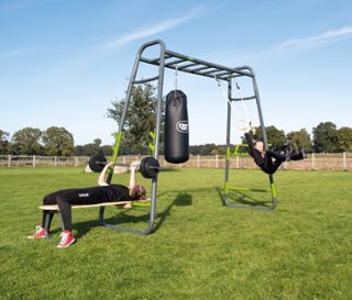 Eine Frau trainiert mit einer Hantel auf dem EXIT Outdoor-Fitnessgerät »MB230«, während ein Mädchen an Ringen schwingt.