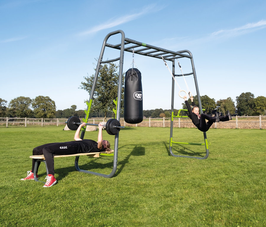 Eine Frau trainiert mit einer Hantel auf dem EXIT Outdoor-Fitnessgerät »MB230«, während ein Mädchen an Ringen schwingt.