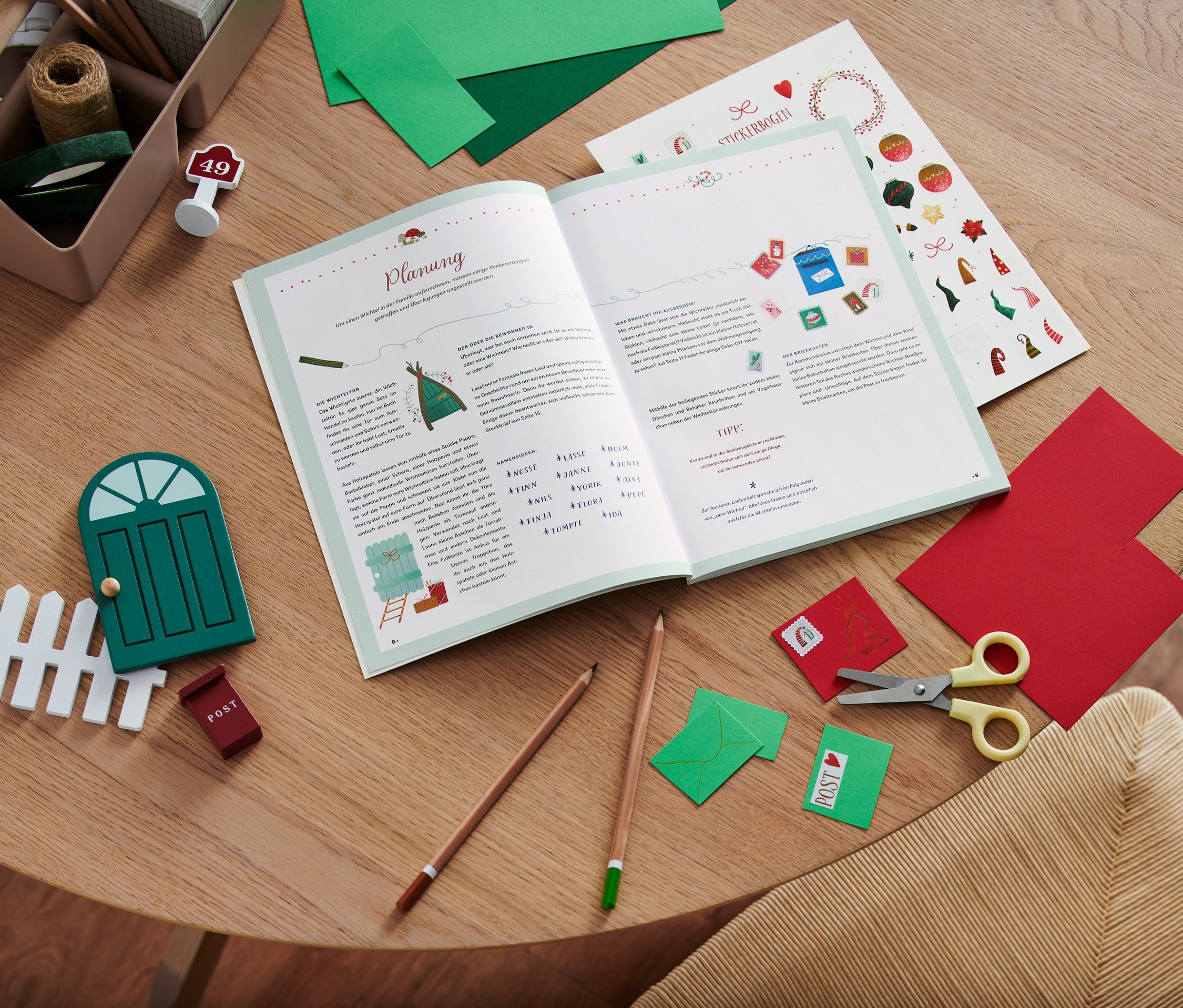 Auf einem Holztisch liegen ein offenes Buch, eine Schere, Buntstifte, bunte Papiere und Dekoartikel, inklusive das Buch »Hurra, ein Wichtel zieht ein!«.