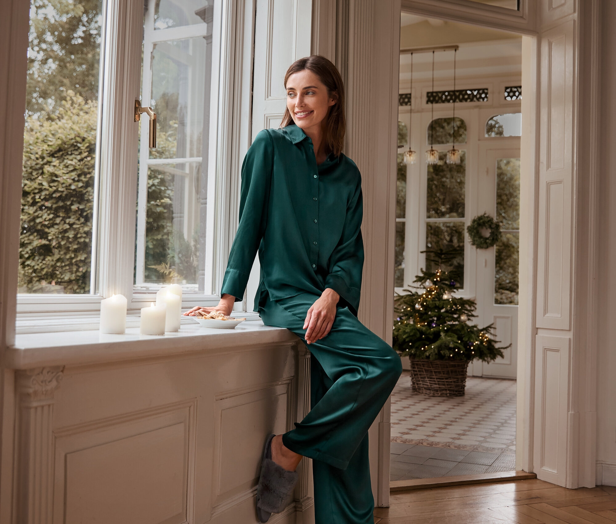 Eine Frau sitzt in grüner Seidenpyjamahose, Seidenoberteil und Plüsch-Hausschuhen auf einer Fensterbank.