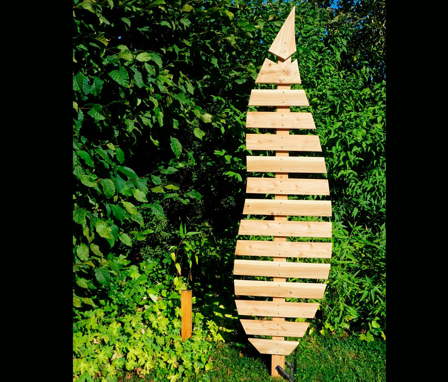 Eine hölzerne Rankhilfe »Bambus« in Blattform steht auf einer Wiese vor Büschen.