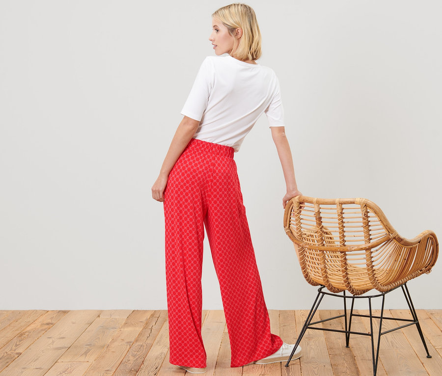 Frau posiert in rot bedruckter Palazzohose und weißem T-Shirt neben einem Rattansessel.