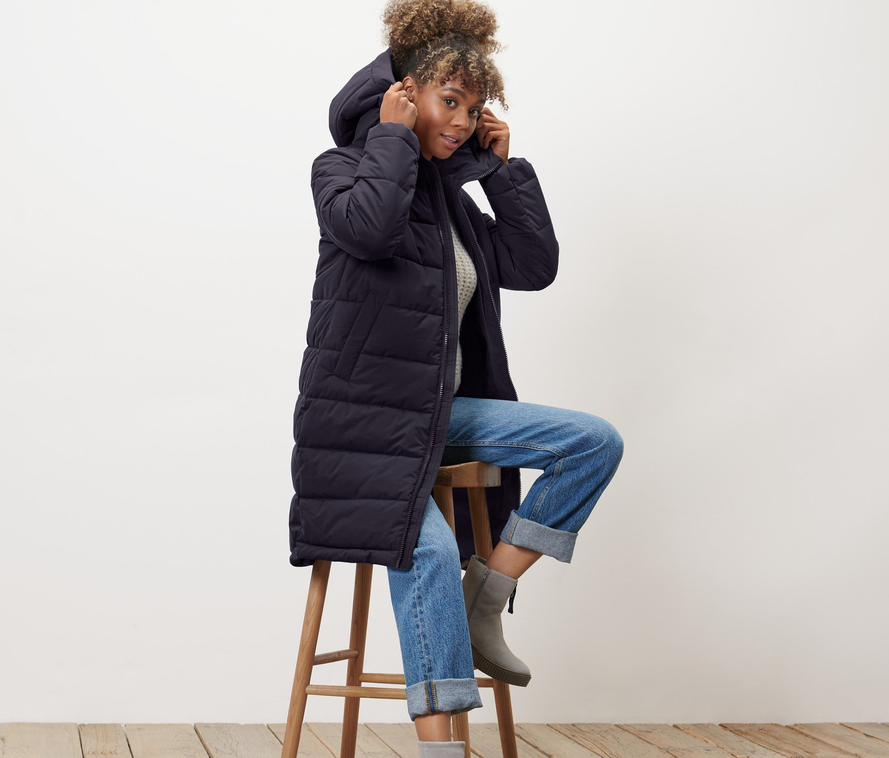 Frau sitzt auf einem Hocker und richtet die Kapuze ihres blauen Steppmantels.