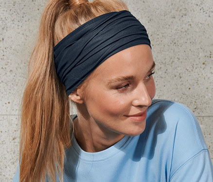 Frau mit blonden, zum Pferdeschwanz gebundenen Haaren trägt ein Multifunktionstuch und ein blaues Oberteil.