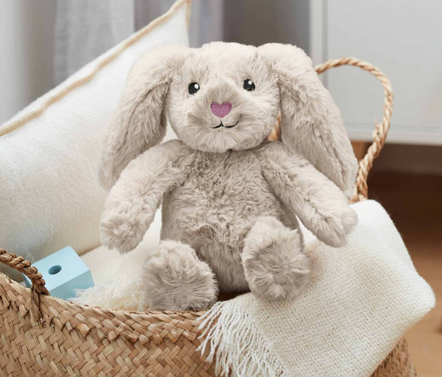 Ein Plüsch-Hase sitzt in einem Weidenkorb mit einem weißen Kissen und einer Decke.