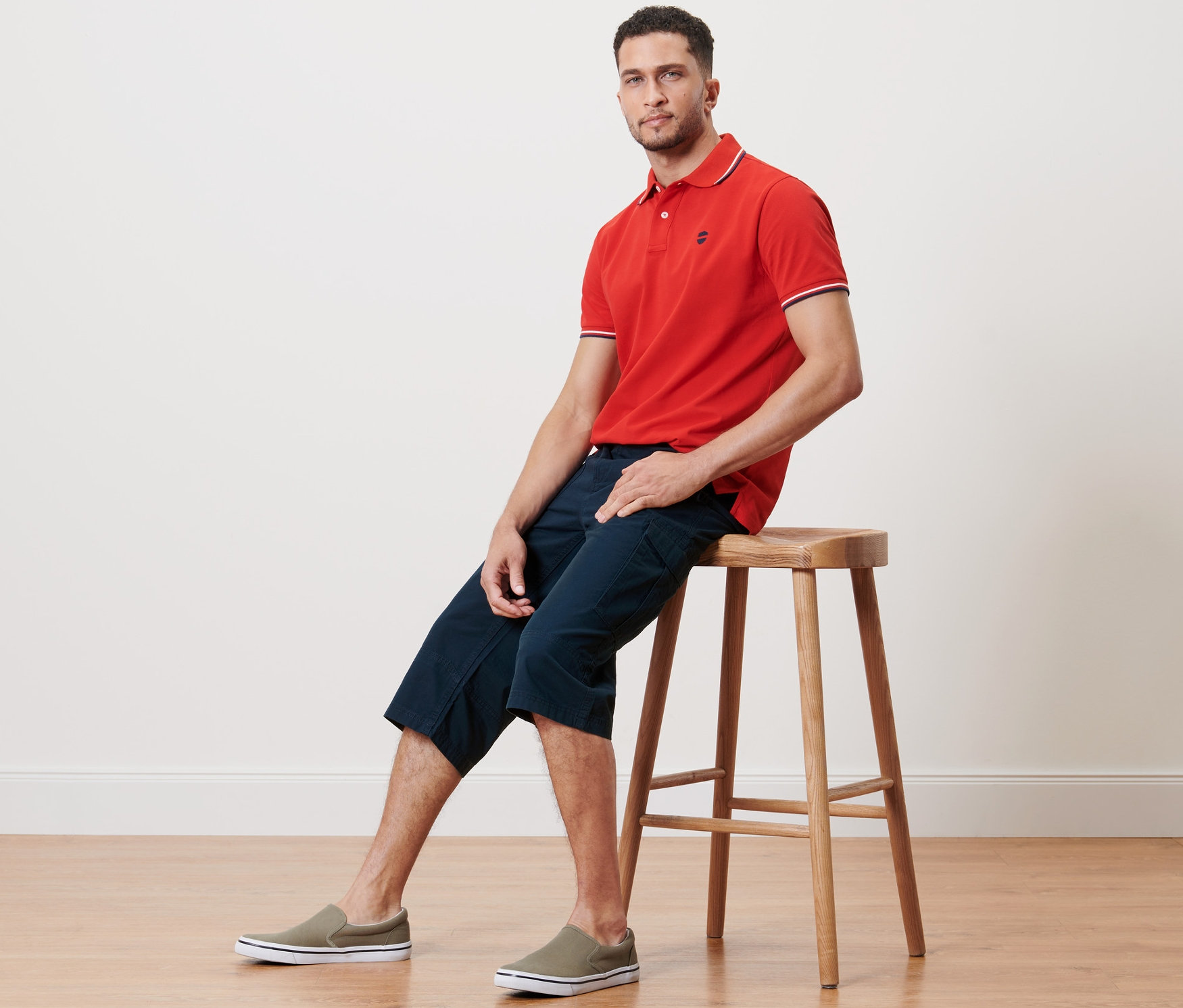 Ein Mann sitzt auf einem Hocker und trägt ein rotes Poloshirt und eine Cargo-Bermuda in 3/4-Länge.