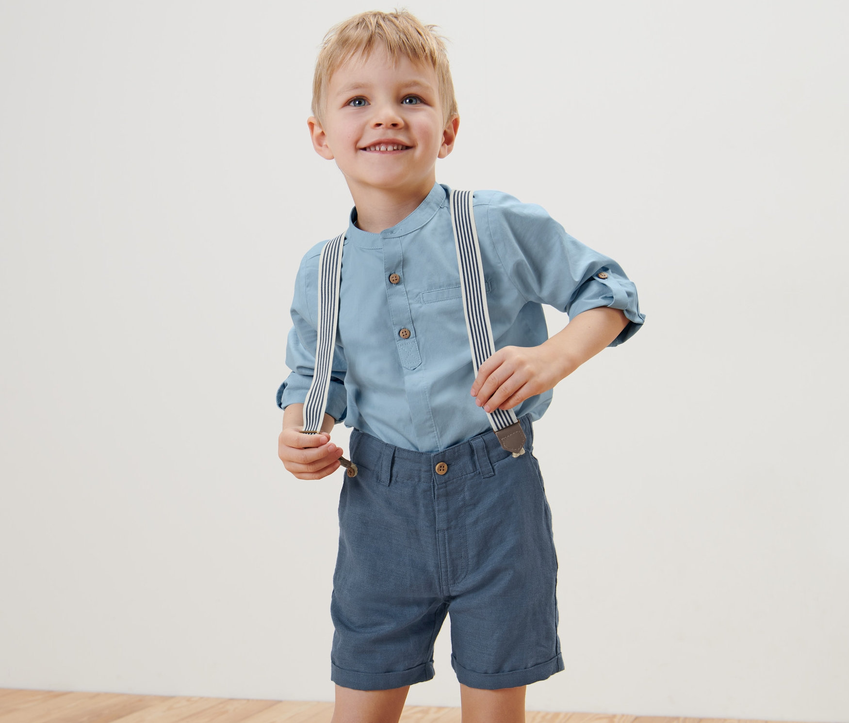 Ein Junge trägt ein blaues Hemd und blaue Kinder-Shorts und Hosenträger und lächelt.