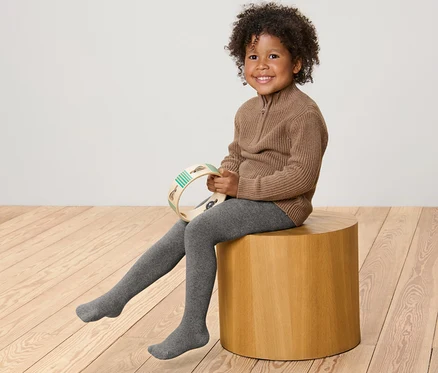Ein lächelndes Kind sitzt auf einem Holzhocker und hält ein Tamburin. Es trägt einen braunen Pullover und eine graumelierte 2 Kinder-Thermostrumpfhosen, meliert.
