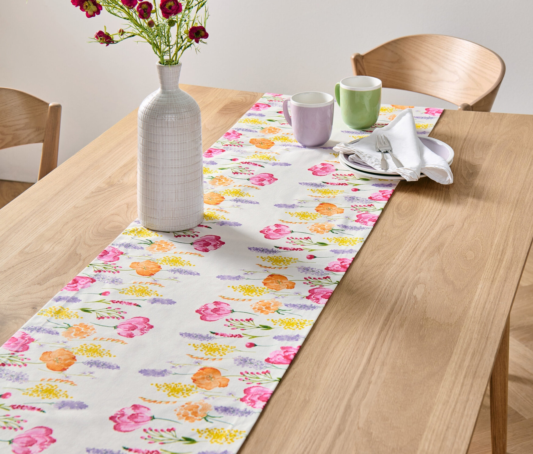 Auf einem Holztisch liegt ein weißer Tischläufer mit Blumendruck. Auf dem Läufer stehen eine Vase mit Blumen und zwei Tassen.