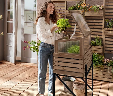 Eine Frau hält Salat neben einem Hochbeet mit Frühbeetaufsatz »Elin«. Im Hintergrund befinden sich ein kleiner Gartenschrank »Tinus« und ein beidseitig bepflanzbarer Sichtschutz »Tinus«.