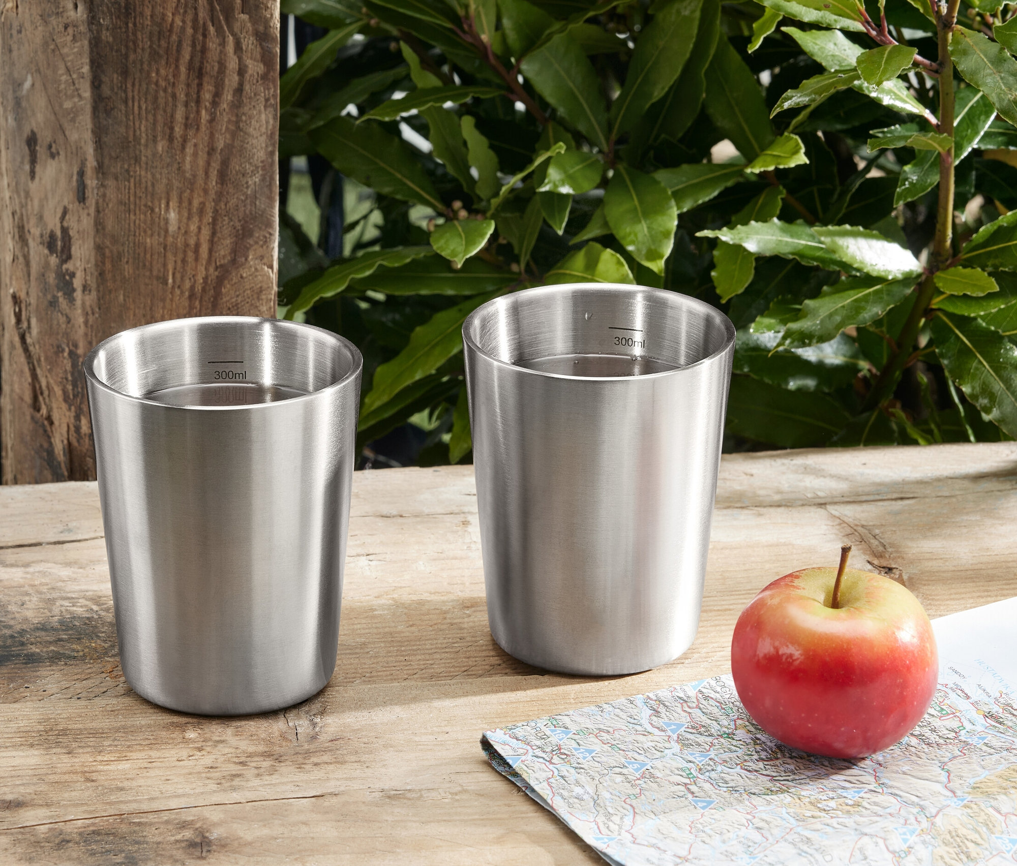 Zwei Camping-Becher aus Edelstahl stehen neben einem Apfel und einer Landkarte auf einem Holztisch.