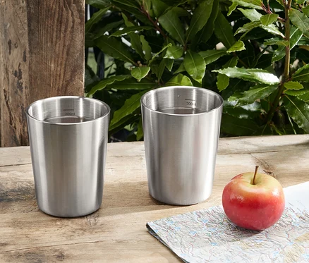 Zwei Camping-Becher aus Edelstahl stehen neben einem Apfel und einer Landkarte auf einem Holztisch.