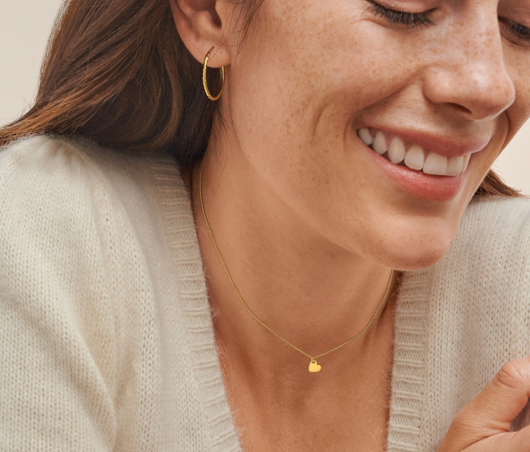 Detailaufnahme einer jungen Frau mit braunen Haaren, die eine Gold-Kette mit Herzanhänger und goldene Ohrringe trägt.