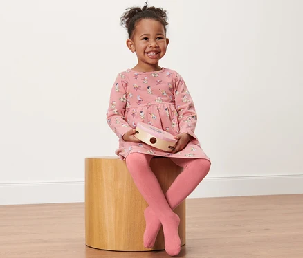 Lächelndes kleines Mädchen sitzt auf einem Holzhocker und hält ein Tamburin. Sie trägt ein rosa Kleid mit Print und 2 Kleinkind-Thermostrumpfhosen, Vogel.