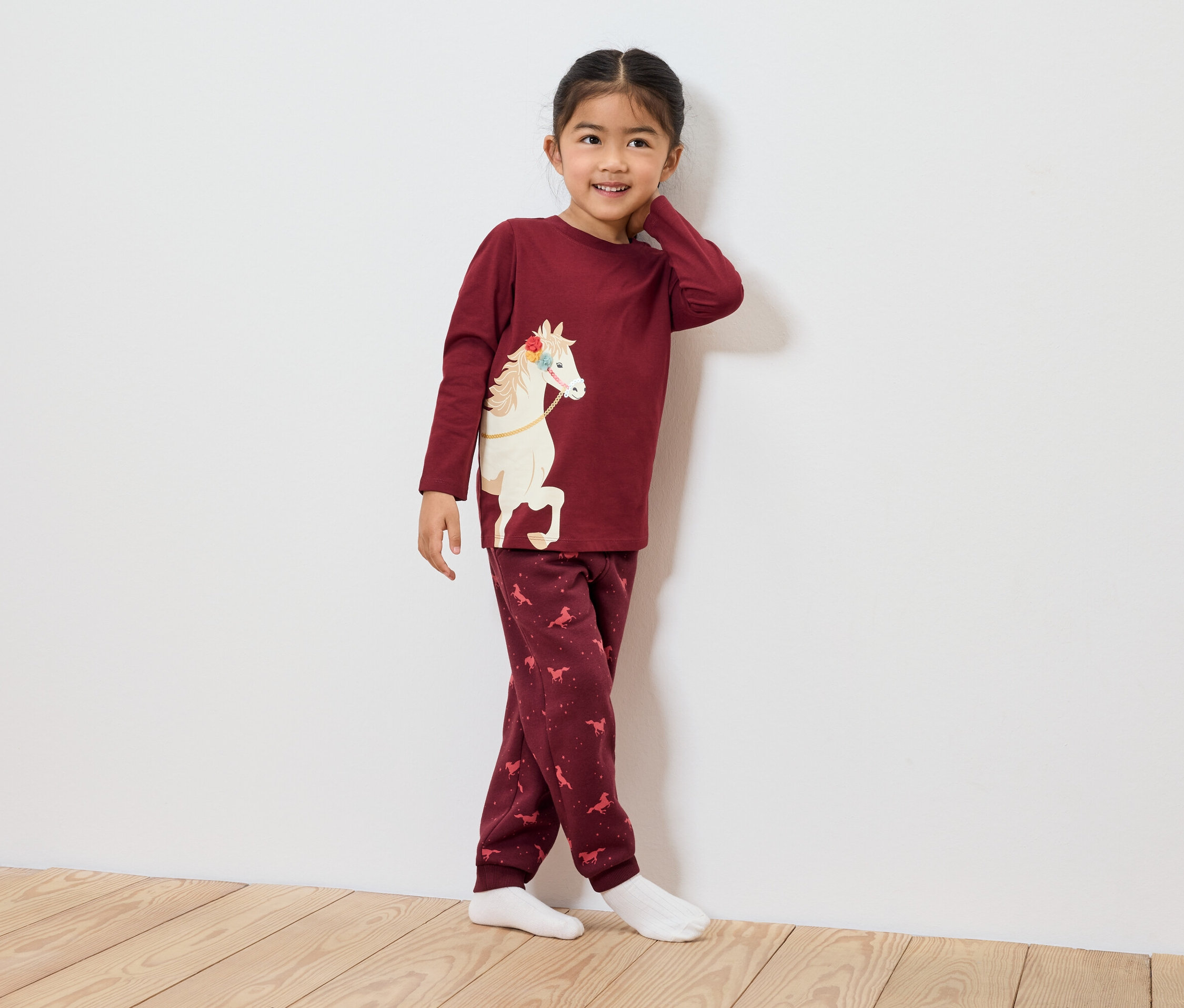 Ein Mädchen im bordeauxfarbenen Pyjama mit Pferdemotiv steht an einer Wand.