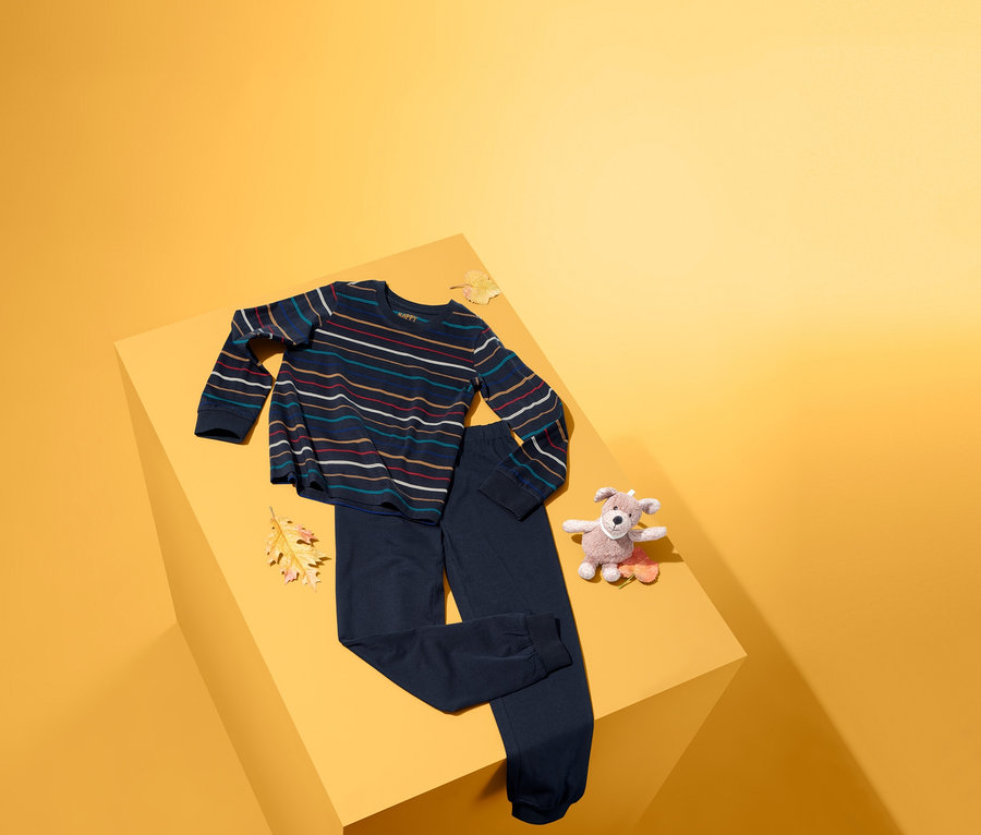 Auf einer gelben Oberfläche liegen ein Pyjama mit einem gestreiften Oberteil und einer dunkelblauen Hose sowie ein Plüschhund.