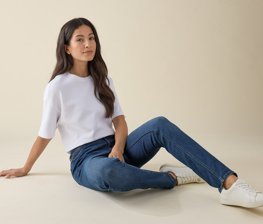 Frau sitzt in Slim-Jeans – Fit »Emma«, Mid Blue Denim, weißem T-Shirt und Turnschuhen am Boden.